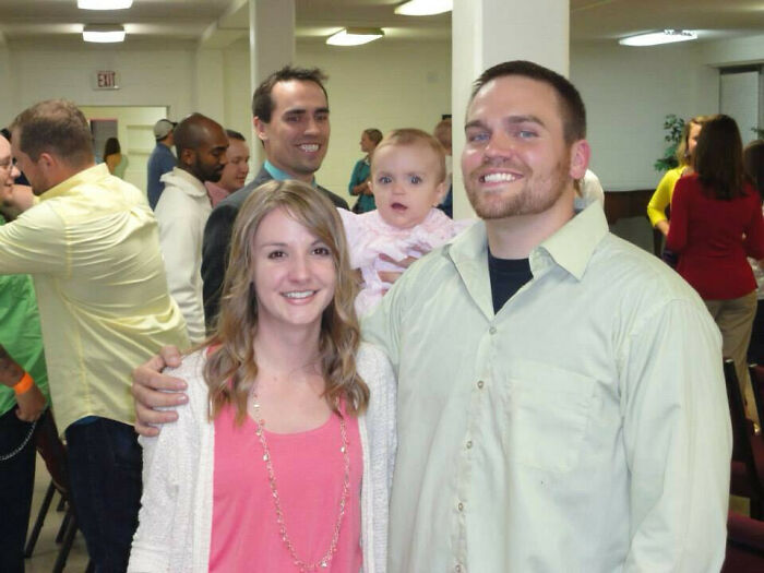 Family posing for a photo at a gathering with a baby photobombing in the background, capturing an epic photobomb moment.