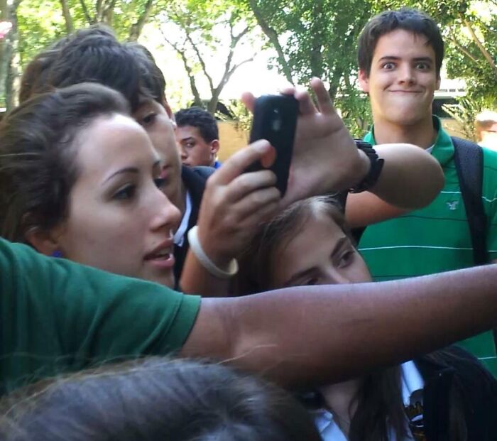 Group taking a selfie outdoors with a person photobombing in the background wearing a green shirt and making a funny face.