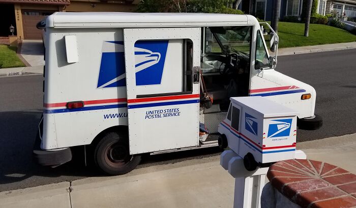 White USPS mail truck parked on a suburban street next to a creative mailbox shaped like a mini postal vehicle. White USPS mail truck parked on a suburban street next to a creative mailbox shaped like a mini postal vehicle.