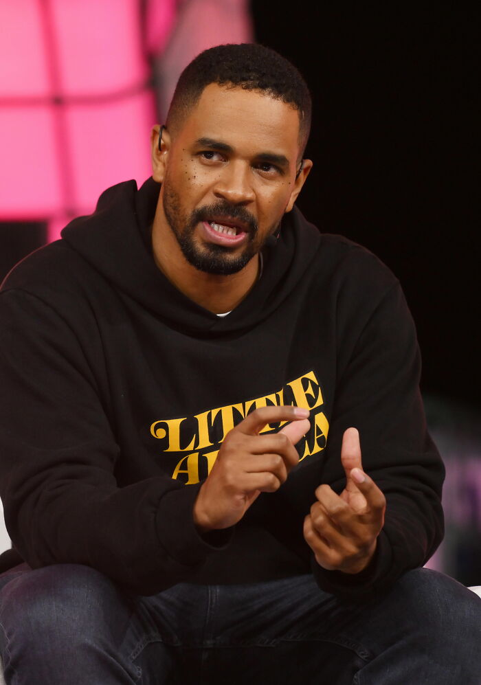 Damon Wayans Jr. speaking during an interview, wearing a black hoodie with yellow text on stage.