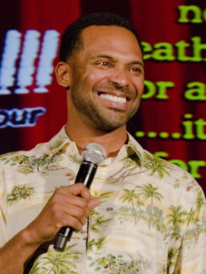 Mike Epps smiling on stage wearing a patterned shirt and holding a microphone during a comedy performance.