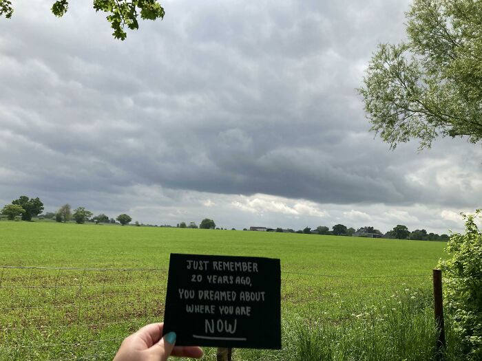 Hand holding a sign with motivational quote in a green field under cloudy sky, inspiring big and small dreams realized.