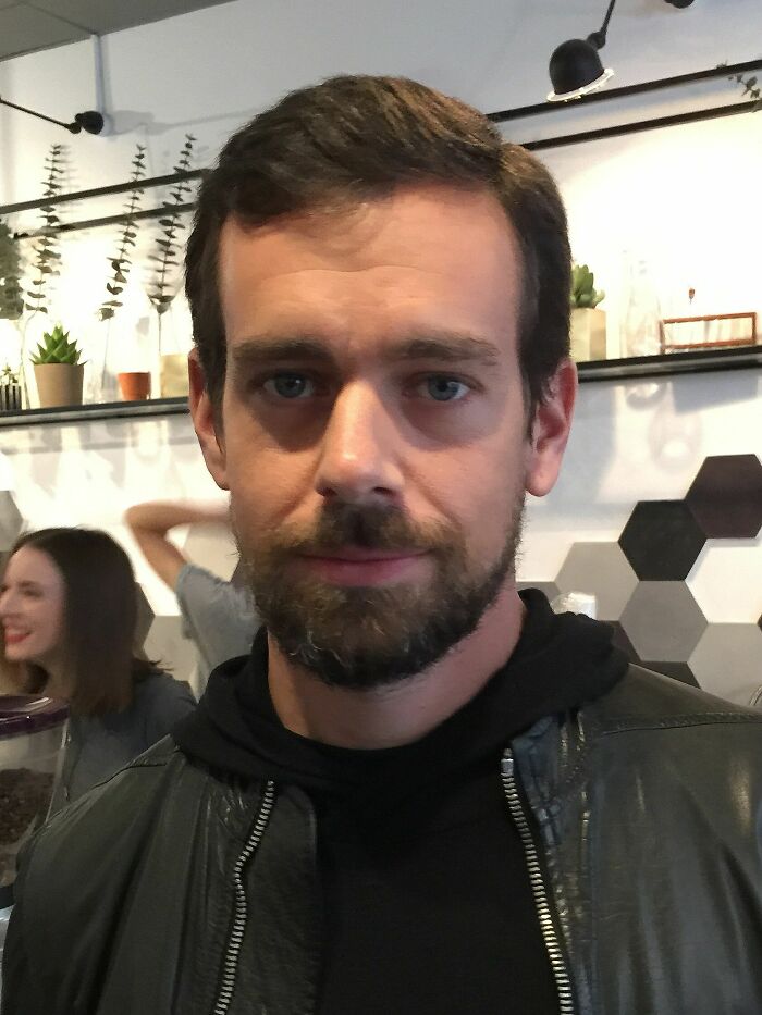 Jack Dorsey with a beard and leather jacket in a casual indoor setting with plants and hexagonal wall decor.