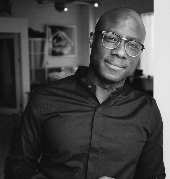 Barry Jenkins wearing glasses and a dark shirt, smiling slightly in a softly lit indoor setting.