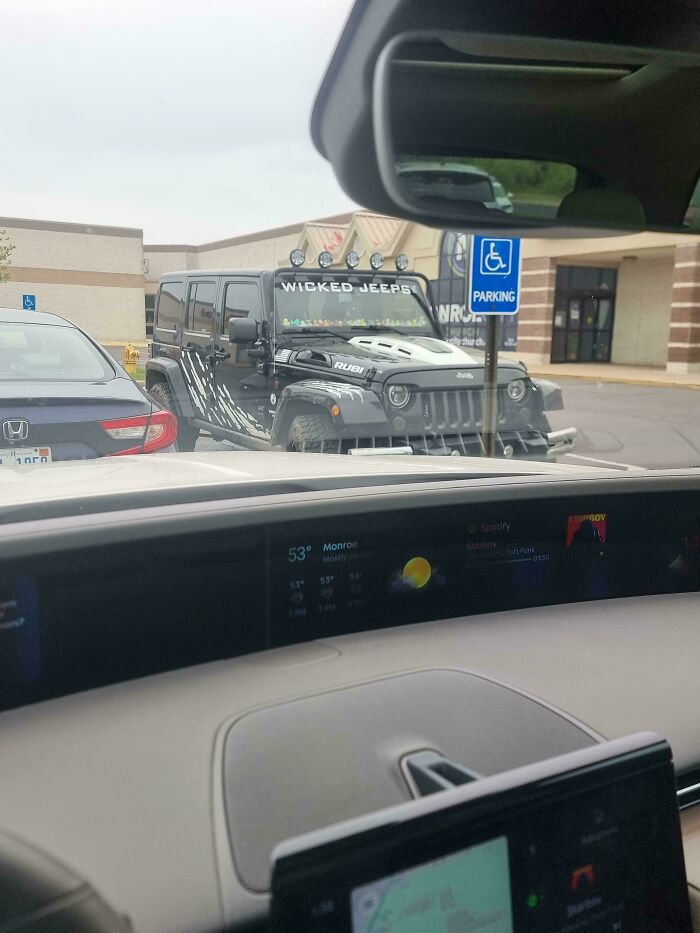 Black Jeep with off-road modifications and "Wicked Jeeps" decal parked improperly in front of a handicap parking sign.