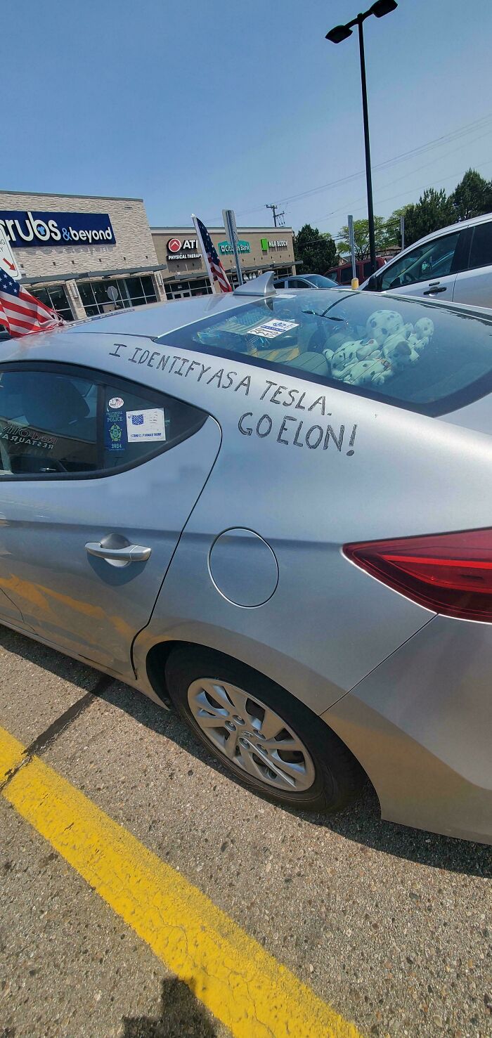 Silver car with handwritten message claiming to be a Tesla, showcasing stupidity on wheels in a parking lot.