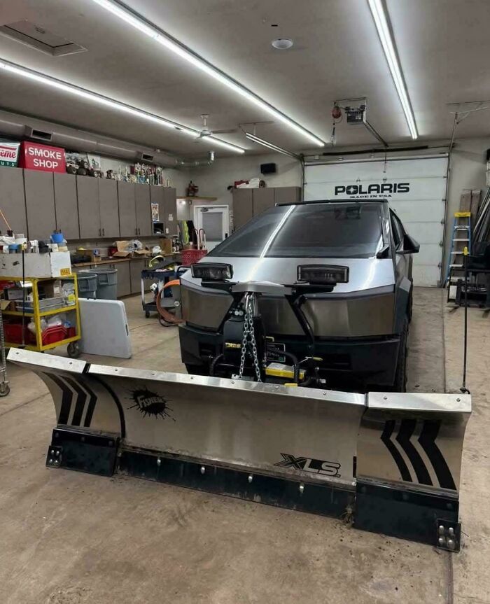 Tesla Cybertruck with an attached large snow plow inside a garage, an example of car modifications gone wrong.