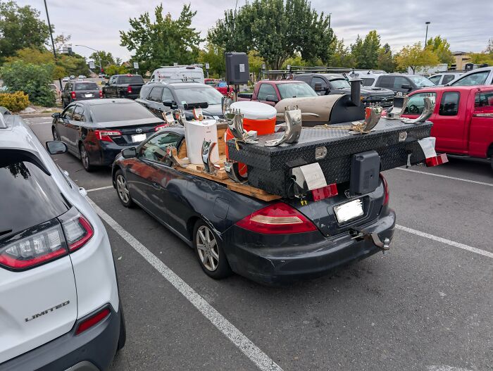 Car modifications gone wrong with an overloaded sedan featuring random objects and parts in a public parking lot.