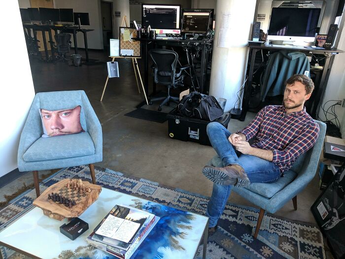 Man sitting in an office chair beside a humorous face pillow on another chair, showcasing employees jokes and pranks at work.