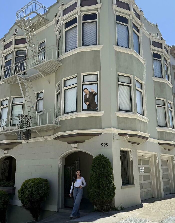 Young woman standing outside a residential building with a man visible through a window, inspiring big and small dreams.