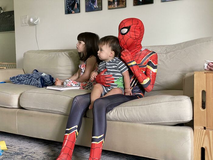 Sibling gratitude captured as a child dressed as Spiderman holds baby brother on a couch next to their sister.