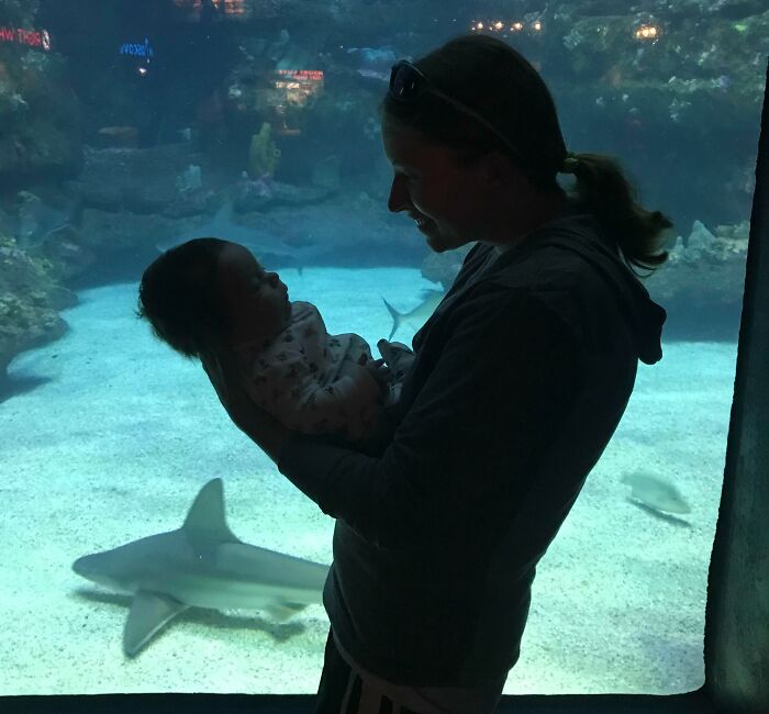 Woman holding baby in front of aquarium with a shark, showing a grateful sibling moment.