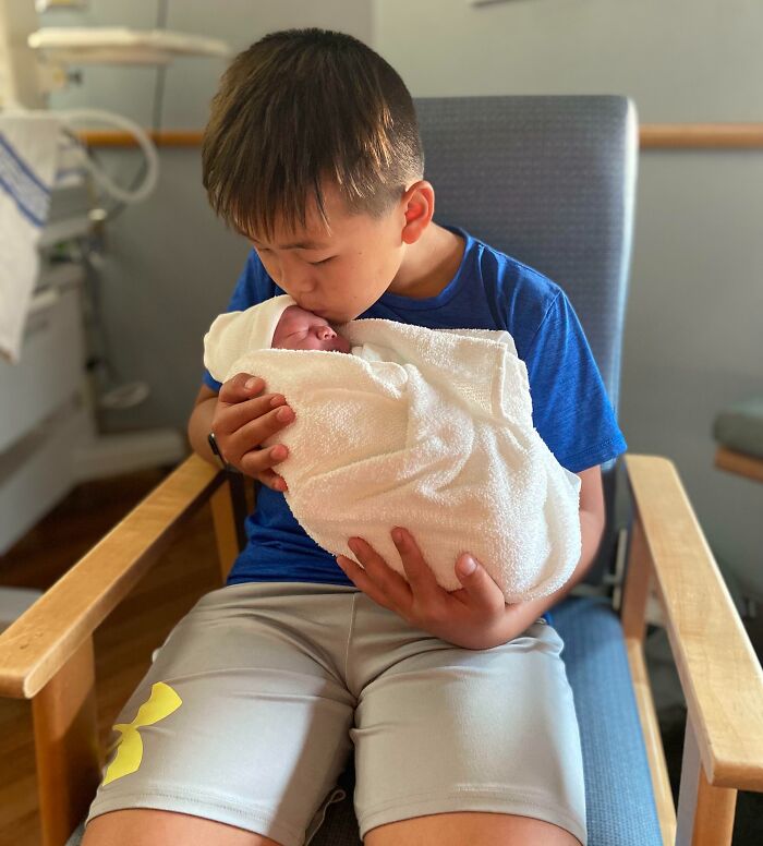 Young boy holding and kissing newborn sibling wrapped in a blanket, showing gratitude and sibling love in a hospital setting.