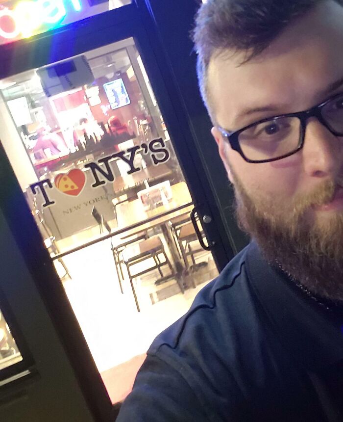 Man with glasses and beard taking selfie outside a pizza restaurant, showcasing dreams turned into reality moment.
