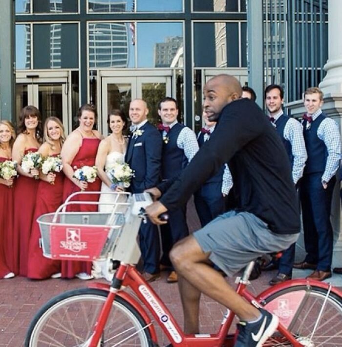 Man riding a bike photobombing a wedding party posing outside a building, creating a funny and epic photobomb moment.