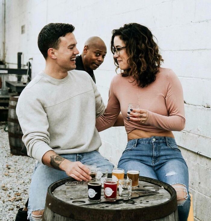 Couple enjoying drinks outdoors with a man photobombing in the background, creating an epic photobomb moment.
