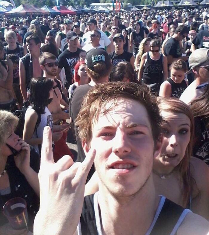 Young man making rock hand sign selfie with unexpected photobomb in crowd at outdoor music festival capturing epic photobombs.
