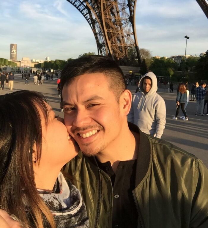 Couple taking a selfie near the Eiffel Tower with a photobombing person in a hoodie in the background.