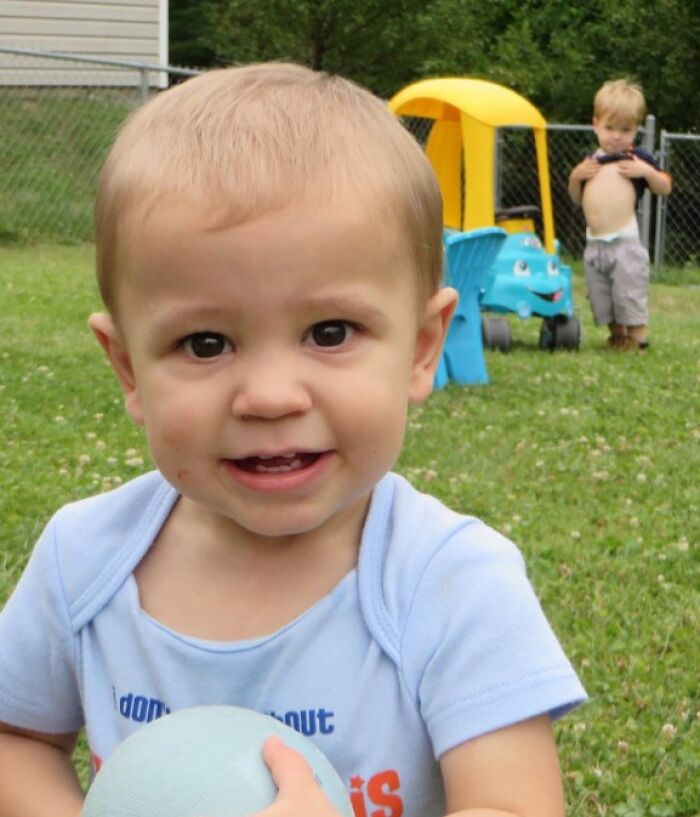 Toddler holding a ball in front with another child photobombing by lifting his shirt in a backyard setting.