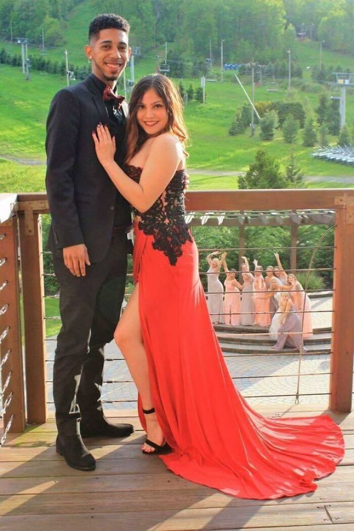 Couple dressed for prom posing on balcony while bridesmaids photobomb with fun gestures in the background.
