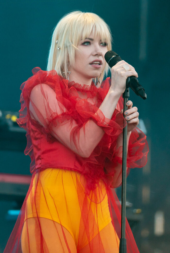 Carly Rae Jepsen performing live on stage wearing a red ruffled top and yellow outfit, holding a microphone.