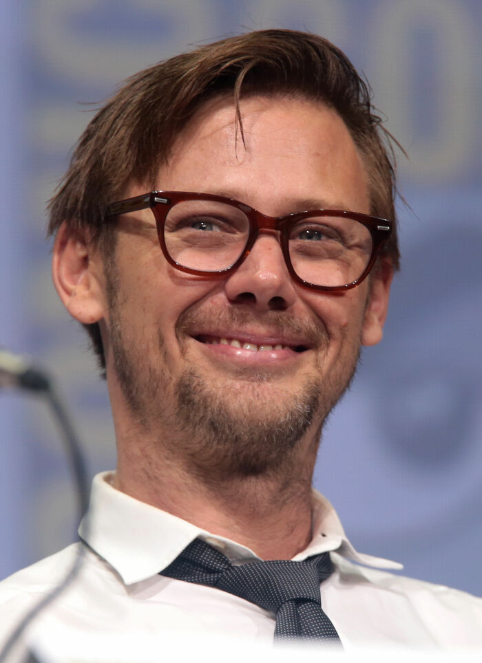 Jimmi Simpson smiling wearing glasses and a white shirt with a dark tie during a public speaking event.