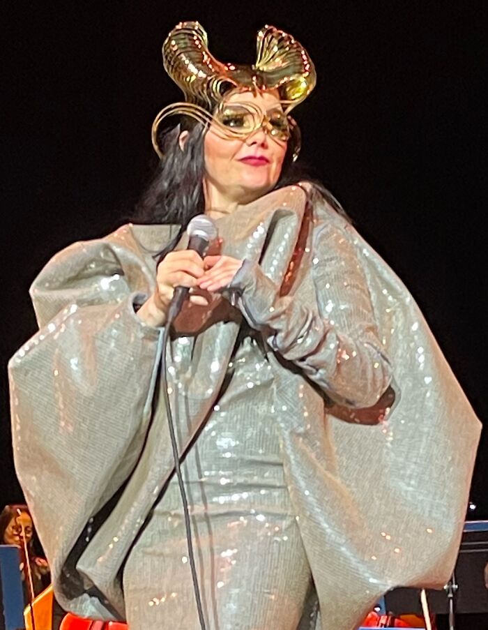 Björk performing live on stage wearing an elaborate metallic costume and holding a microphone during a concert.