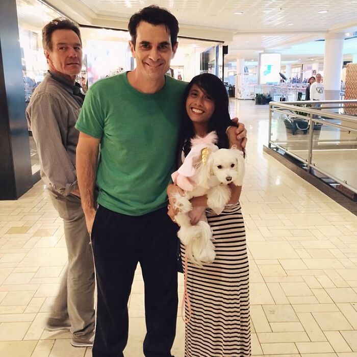 Man and woman smiling with white dog while a man photobombs unexpectedly in a mall hallway.