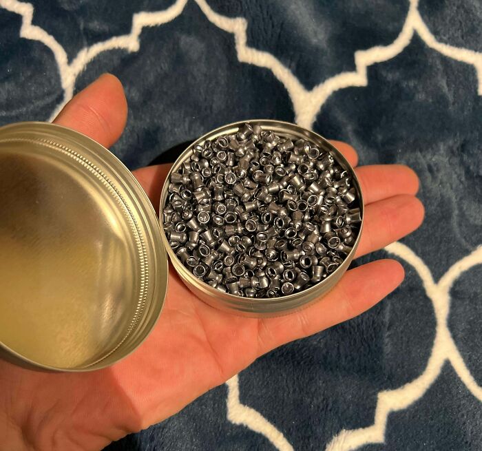 Hand holding an open tin filled with small metal pellets against a patterned dark blue and white background.