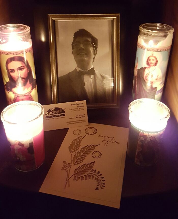 Memorial setup with a framed photo, candles, a sympathy card, and a business card representing employees with jokes and pranks.