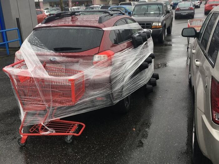 Red car wrapped in plastic with attached shopping carts in a parking lot, a prank by employees who walked out with jokes.