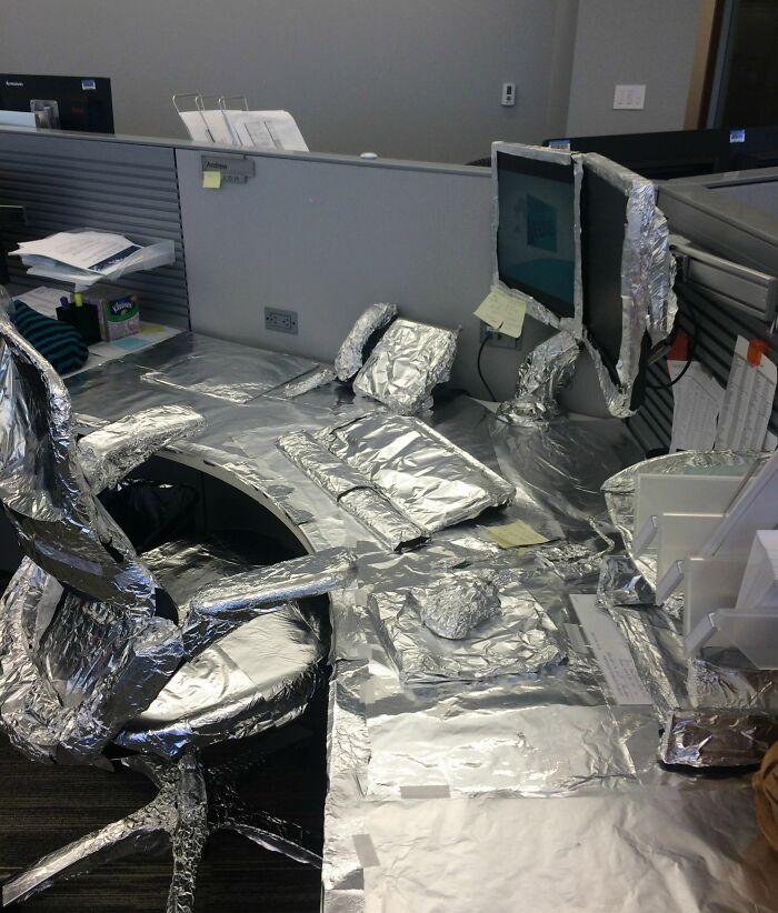 Office desk and chair covered in foil as a prank by employees walking out with jokes and zero regrets.