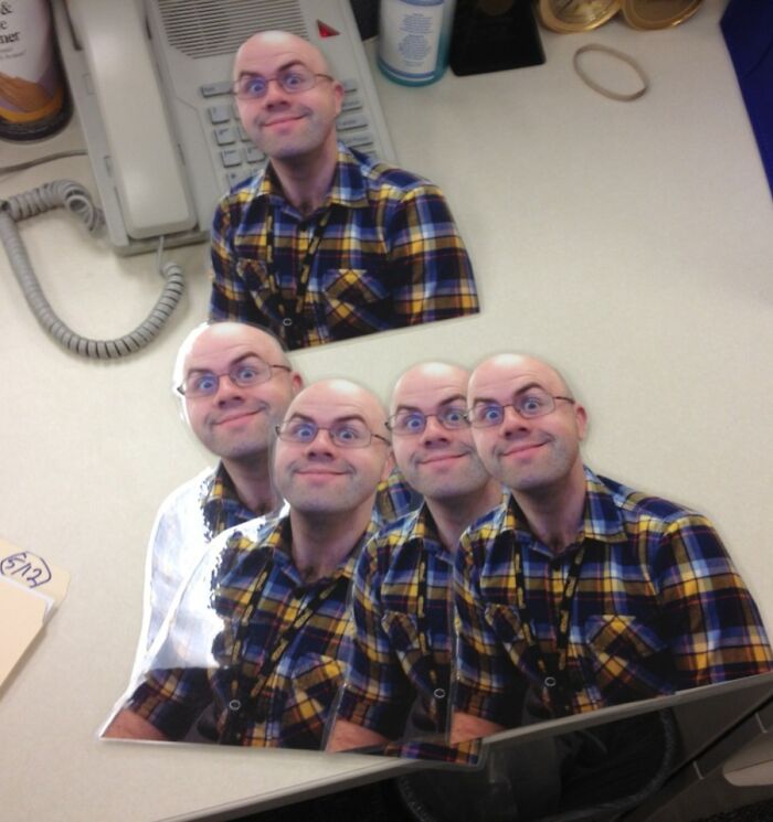 Man in a yellow plaid shirt with multiple cutouts of himself on an office desk, showcasing employee jokes and pranks.