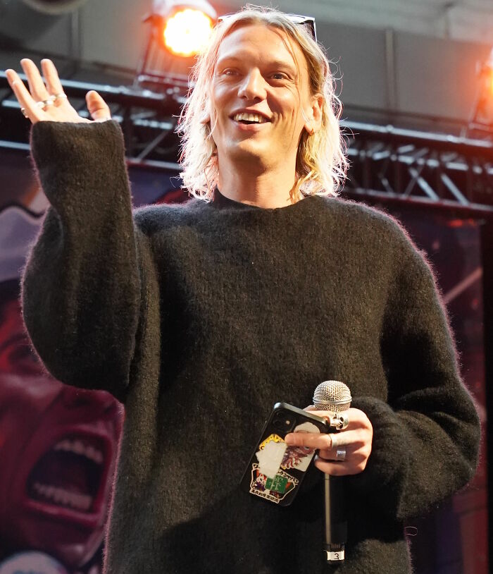Jamie Campbell Bower speaking on stage, holding a microphone and phone, wearing a black sweater under bright lights.
