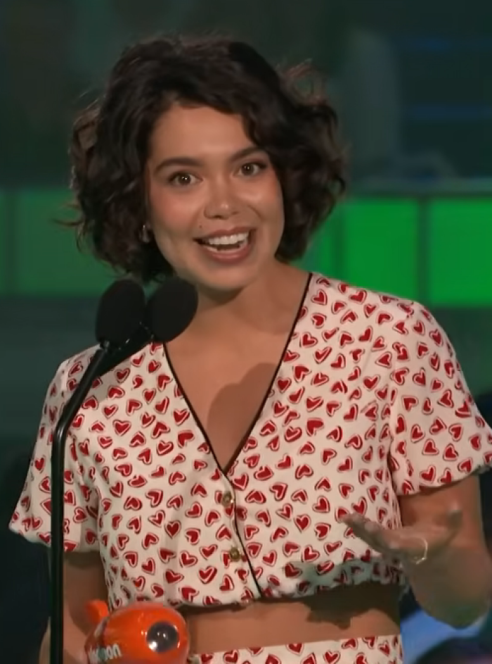 Auliʻi Cravalho speaking at a podium wearing a heart-patterned outfit during a public event.