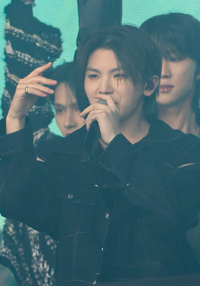 Woozi performing on stage, holding a microphone, wearing a black jacket with a focused expression during a live event.