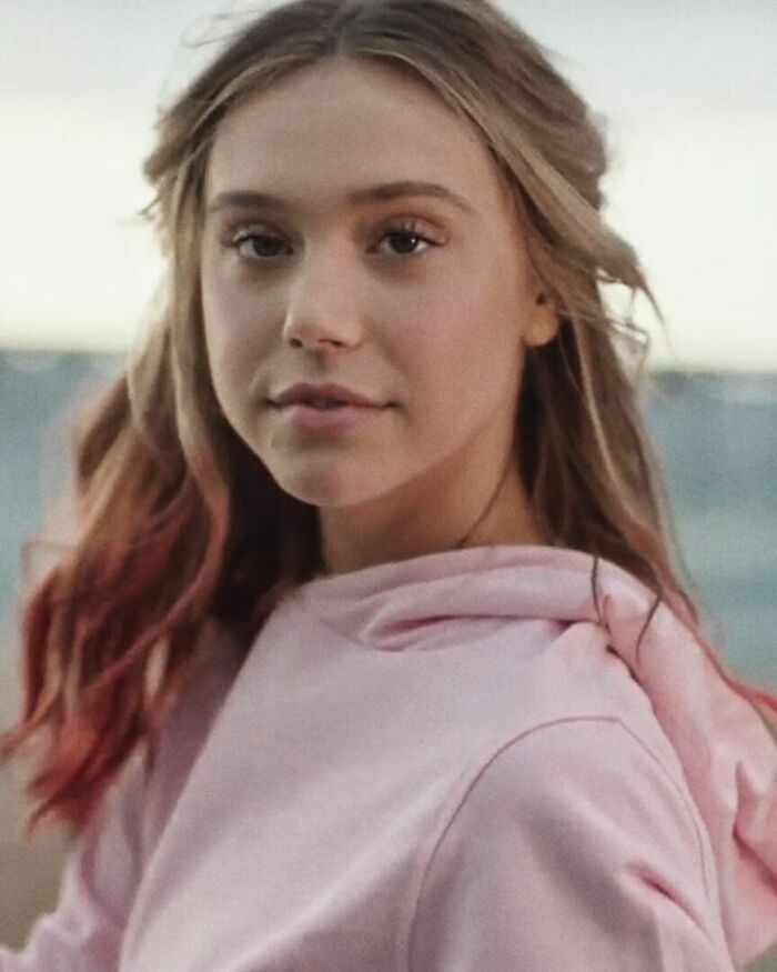 Alexis Ren wearing a pink hoodie, with loose hair, posing outdoors with a blurred background.