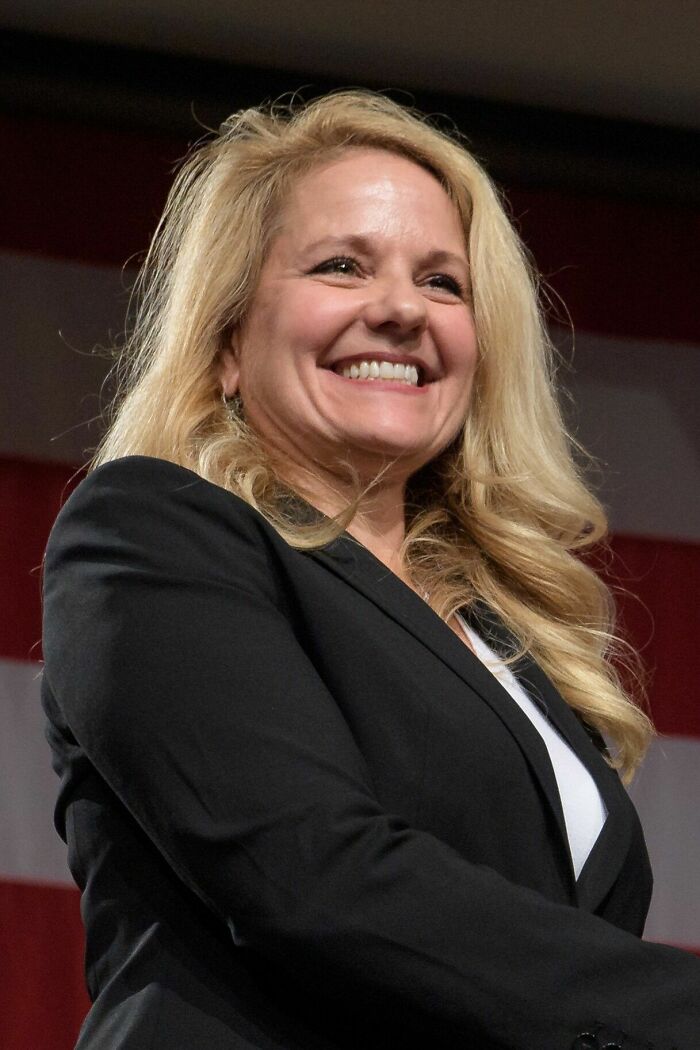 Gwynne Shotwell smiling confidently, wearing a black blazer with a blurred American flag in the background.