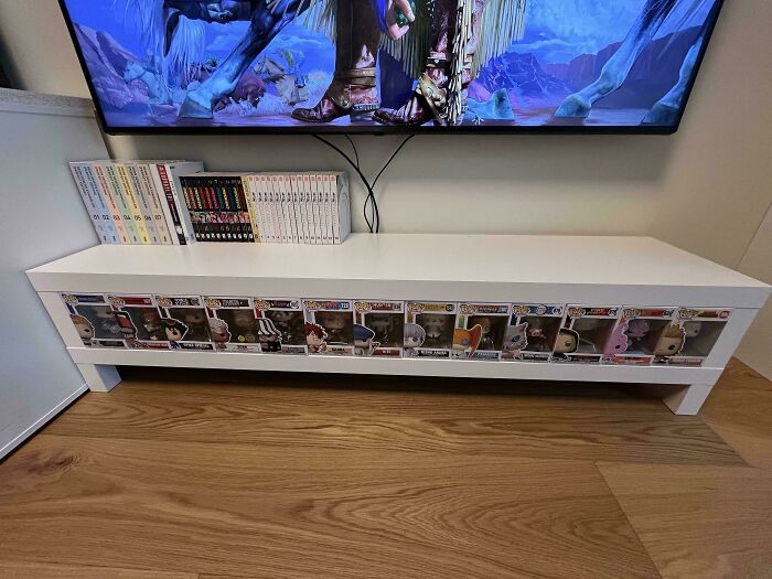 Funko Pop figures perfectly aligned inside a white shelf beneath a TV with manga books arranged on top.