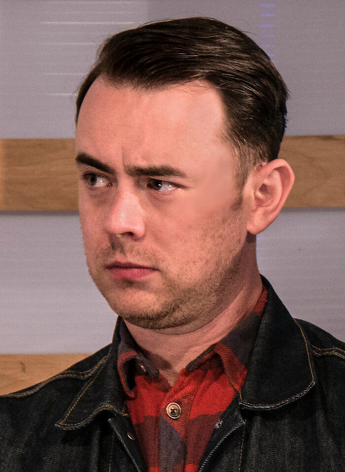 Colin Hanks wearing a denim jacket and red plaid shirt, looking to the side with a serious expression indoors.