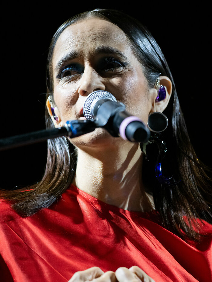 Julieta Venegas singing on stage wearing a red top with earrings and using a microphone during a live performance.