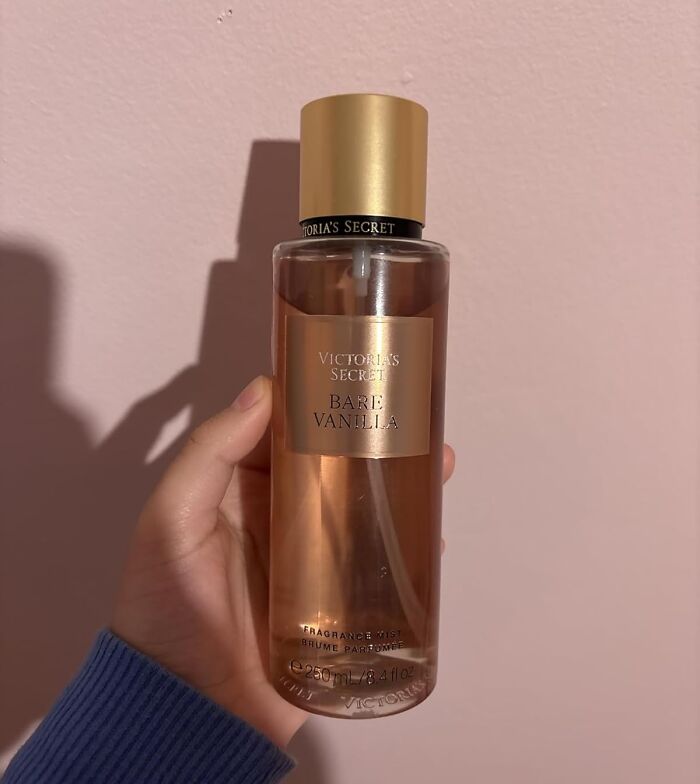 Hand holding a Victoria's Secret Bare Vanilla fragrance mist against a pink wall, a Black Friday stocking stuffer deal.
