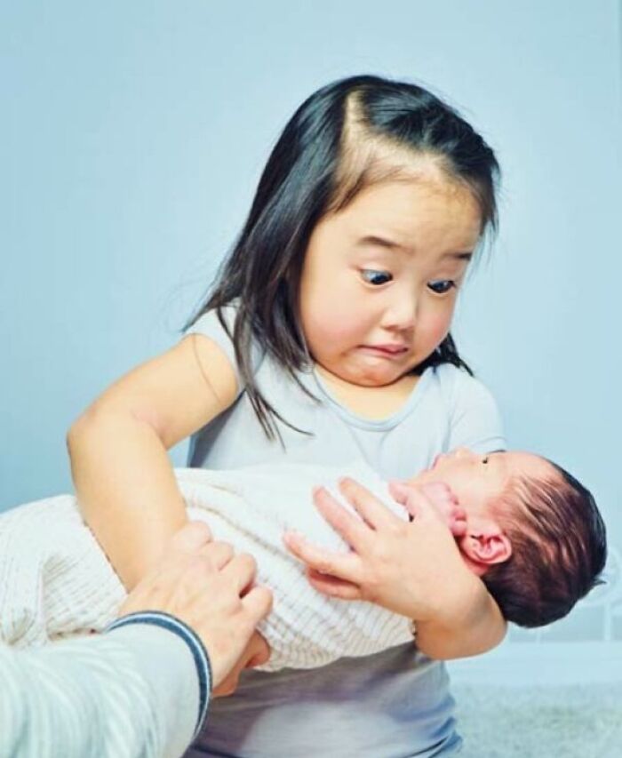 Young girl holding a newborn baby sibling with a surprised expression, capturing a heartfelt sibling moment.