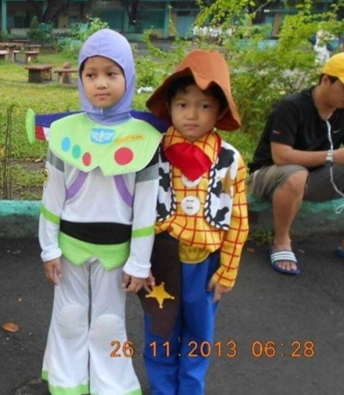 Two young siblings dressed as Buzz Lightyear and Woody, standing outdoors with greenery in the background.