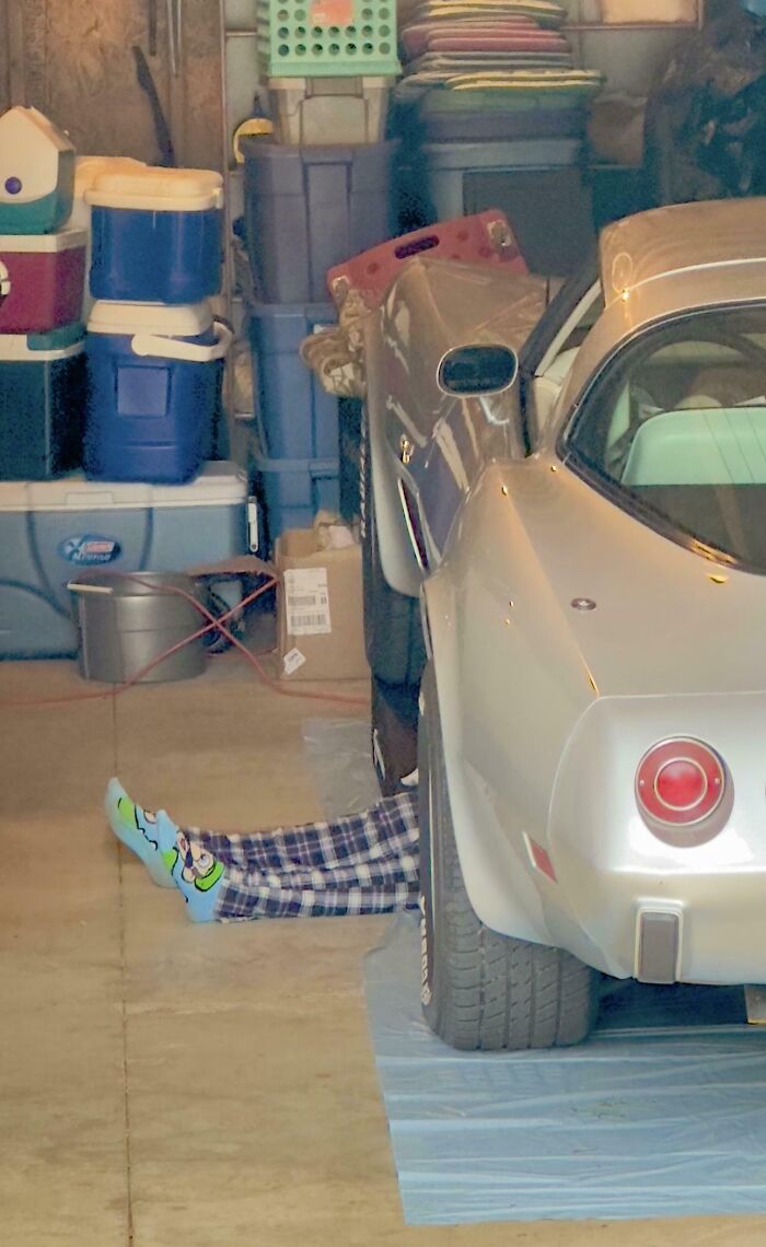 Person wearing colorful socks and plaid pants lies under a silver car in a cluttered garage, showing sibling gratitude.