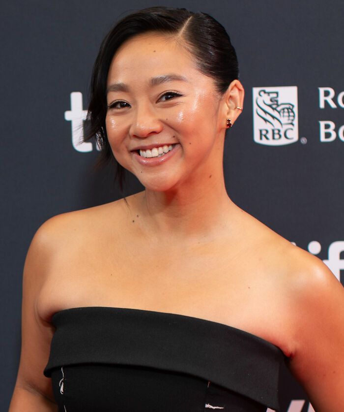 Stephanie Hsu smiling at an event, wearing a black strapless dress with a dark background and logos behind her.