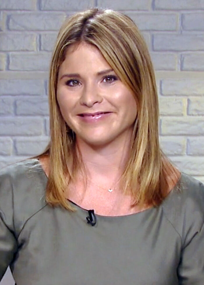 Jenna Bush Hager smiling, wearing a green top and microphone, seated in front of a gray brick wall background.
