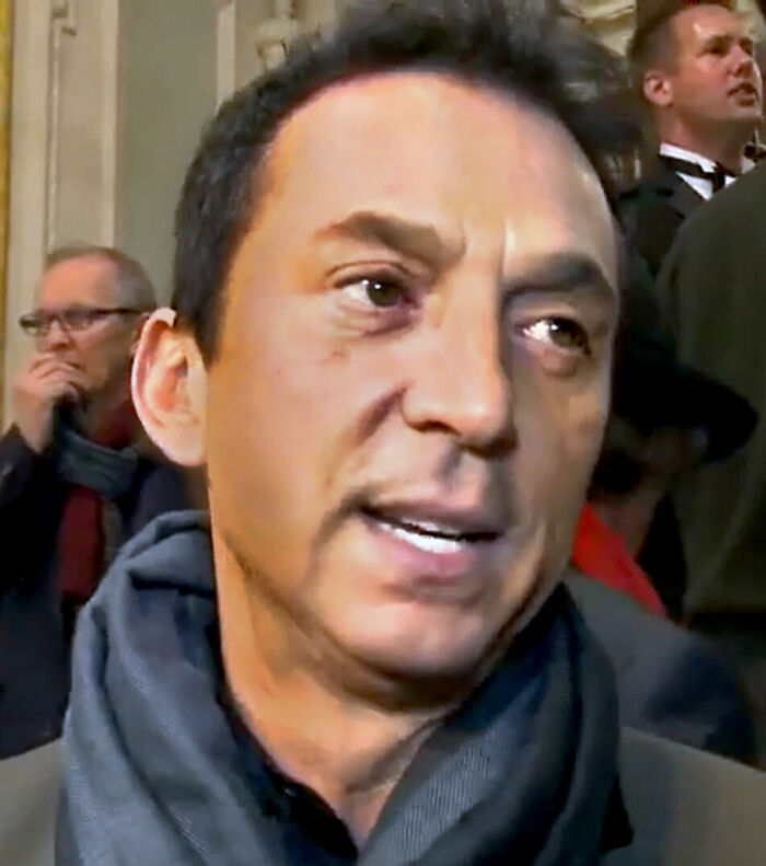 Bruno Tonioli speaking at a public event, wearing a dark scarf and jacket with people in the background.
