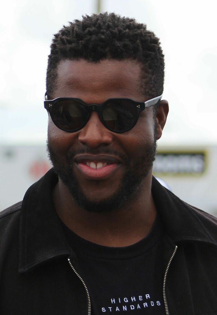 Winston Duke wearing sunglasses and a black jacket, smiling outdoors in a casual setting focusing on career highlights.