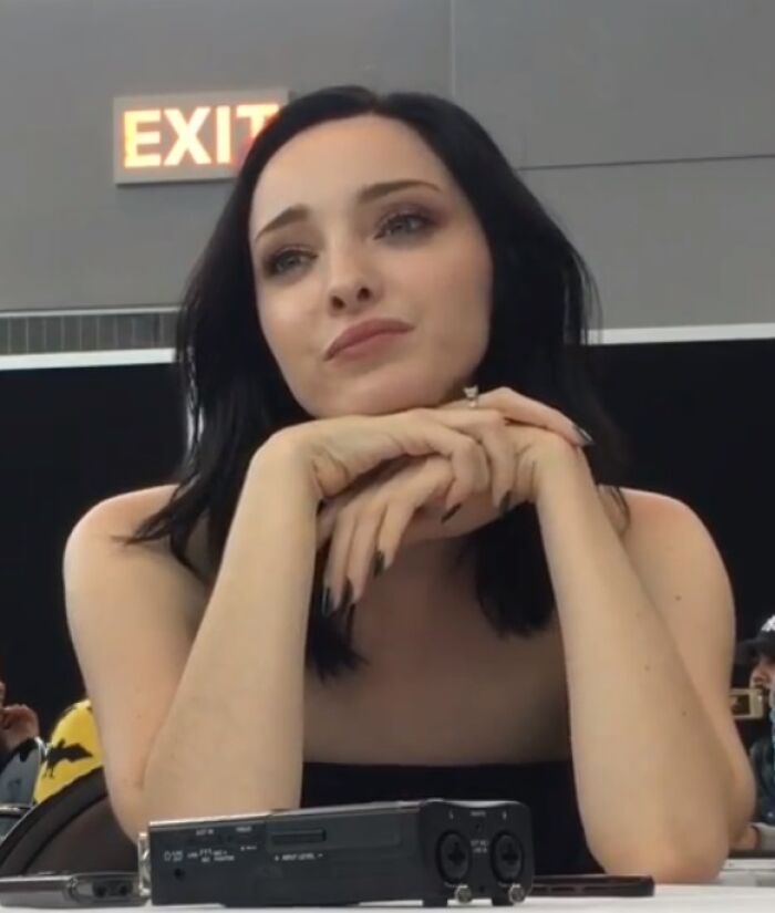 Emma Dumont resting her chin on clasped hands, with dark hair and a thoughtful expression at an indoor event.